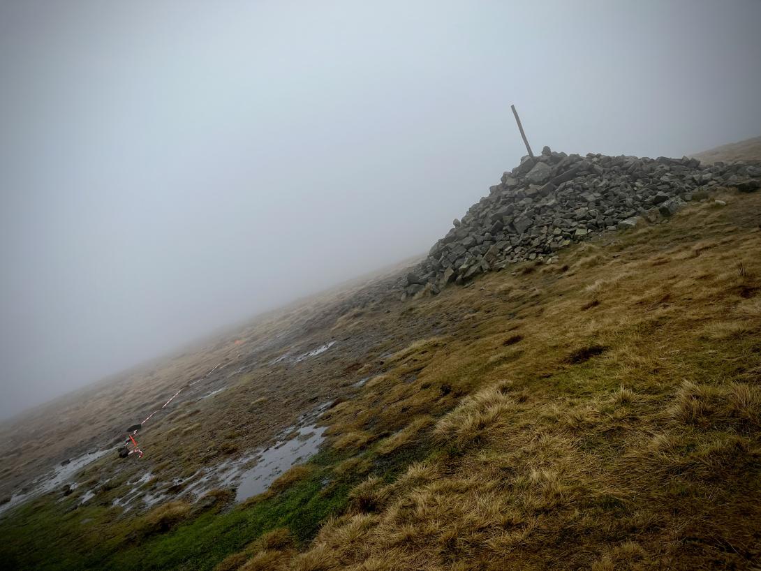 Paddy's Pole: a wooden pole surrounded by rocks and stones on boggy ground on a cloudy day.