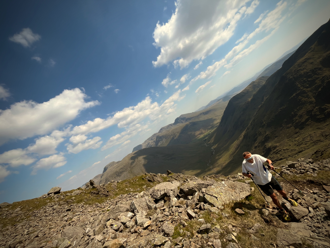 Mike, using hiking poles, making his way up a mountain, blue skies and patchy cloud cover.